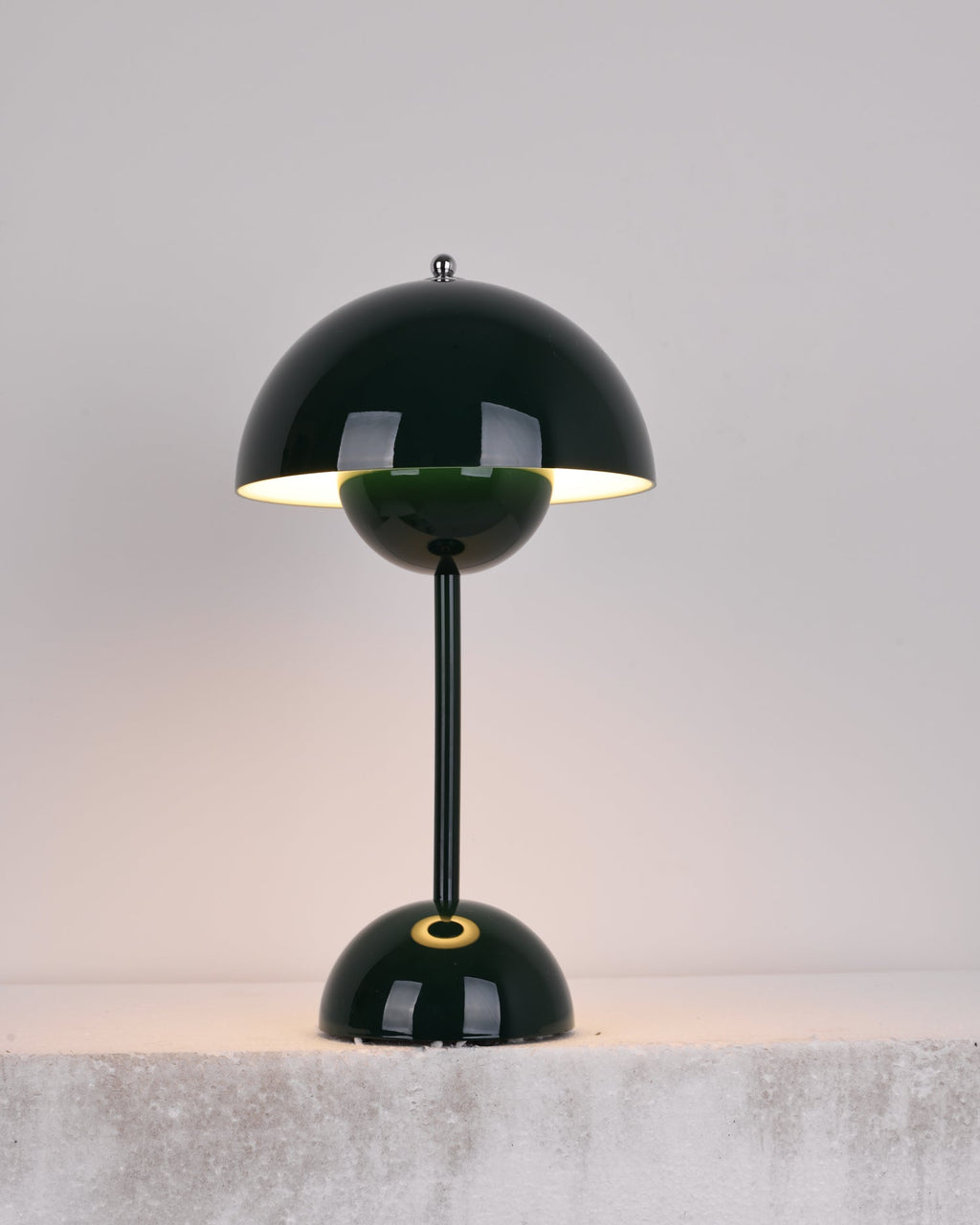 A dark-green Clérachic table lamp lit, with a bright rim under the shade and a gold ring on the base, on a stone ledge.