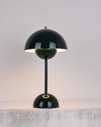 A dark-green Clérachic table lamp lit, with a bright rim under the shade and a gold ring on the base, on a stone ledge.
