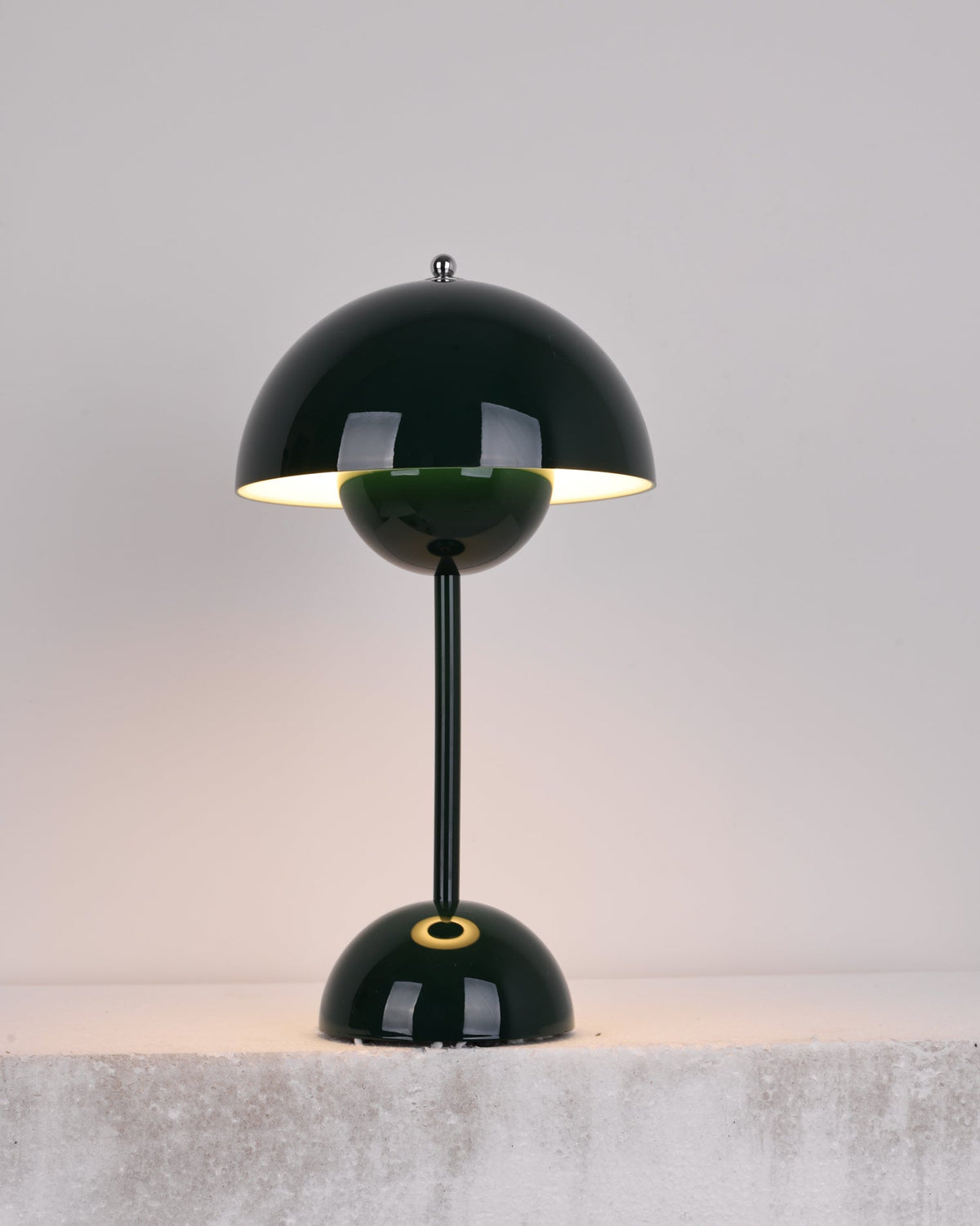 A dark-green Clérachic table lamp lit, with a bright rim under the shade and a gold ring on the base, on a stone ledge.