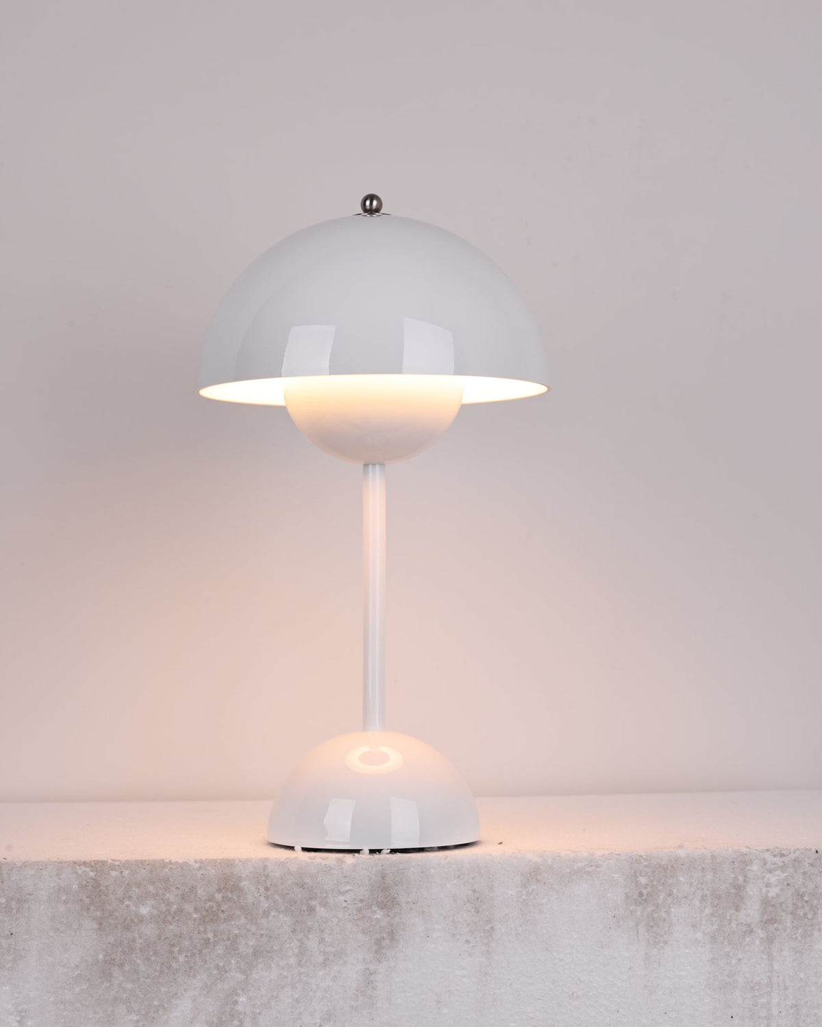 A white Clérachic table lamp lit, with a warm glow below the shade, on a rough pale stone ledge.