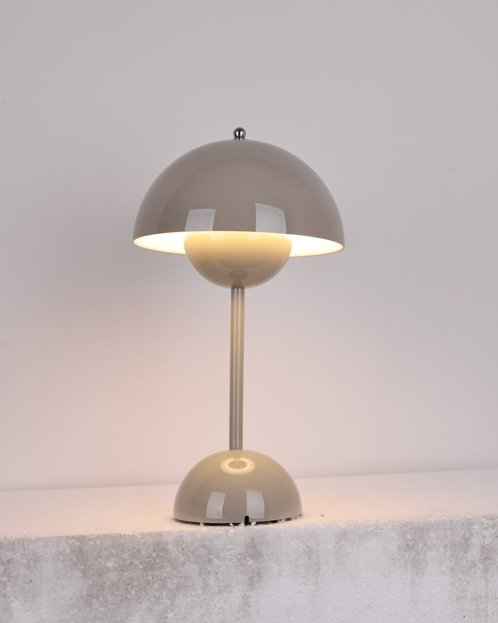 A grey Clérachic table lamp lit, with a warm glow below the shade, on a rough pale stone ledge.