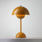 A mustard Clérachic table lamp lit, with a warm glow below the shade and a ring on the base, on a pale ledge.