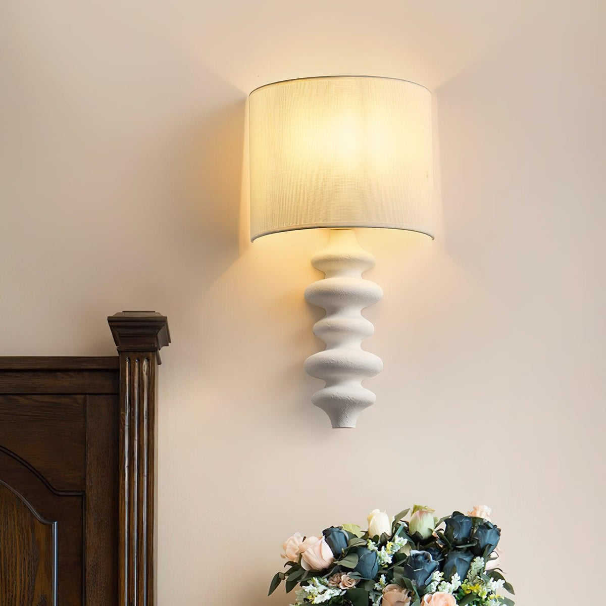 Lit Clérachic wall light with a white linen shade and a white corrugated column, beside a carved wooden bedpost and a bouquet.