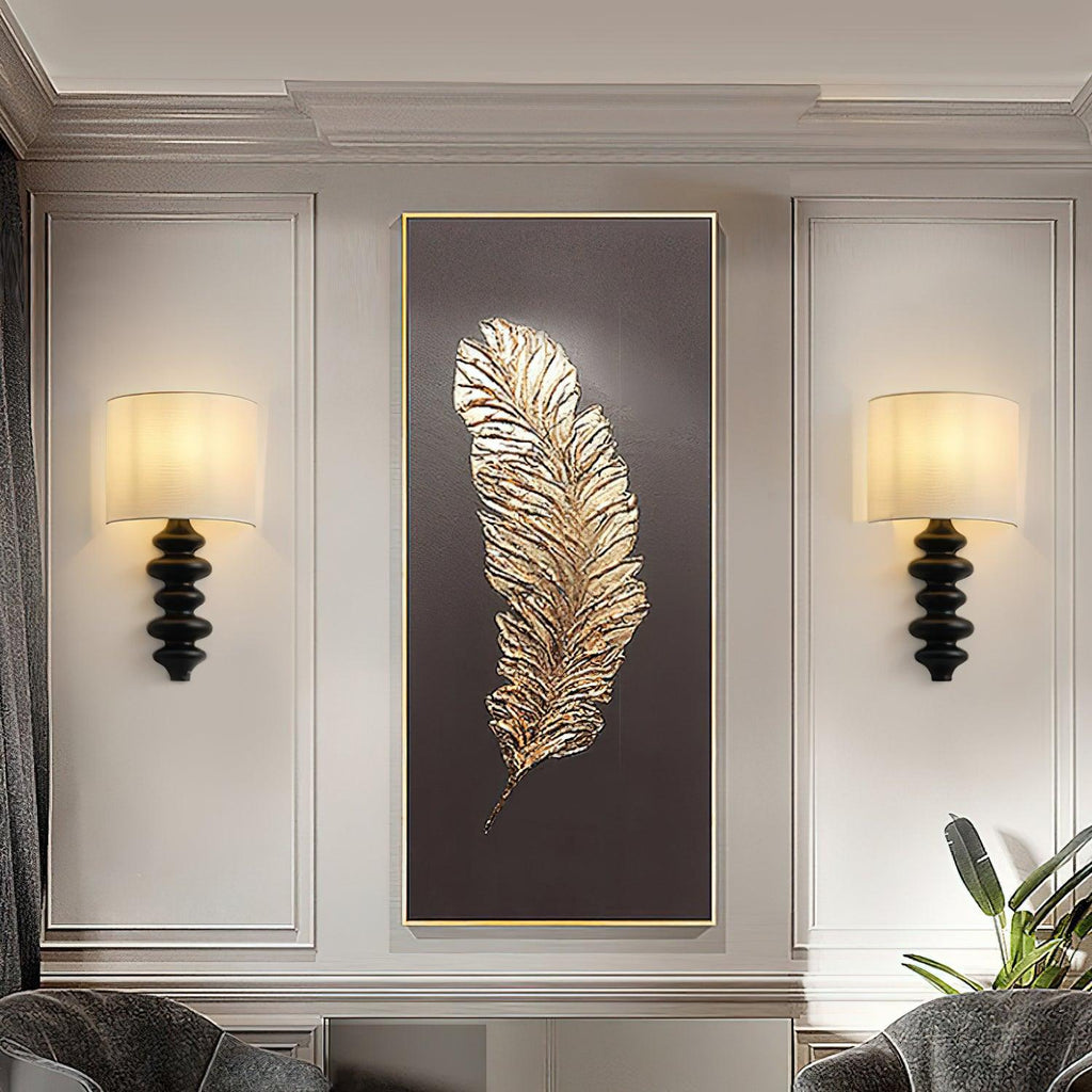 Pair of Clérachic wall lights with black columns and lit linen shades, flanking a large gold feather artwork on a panelled wall.