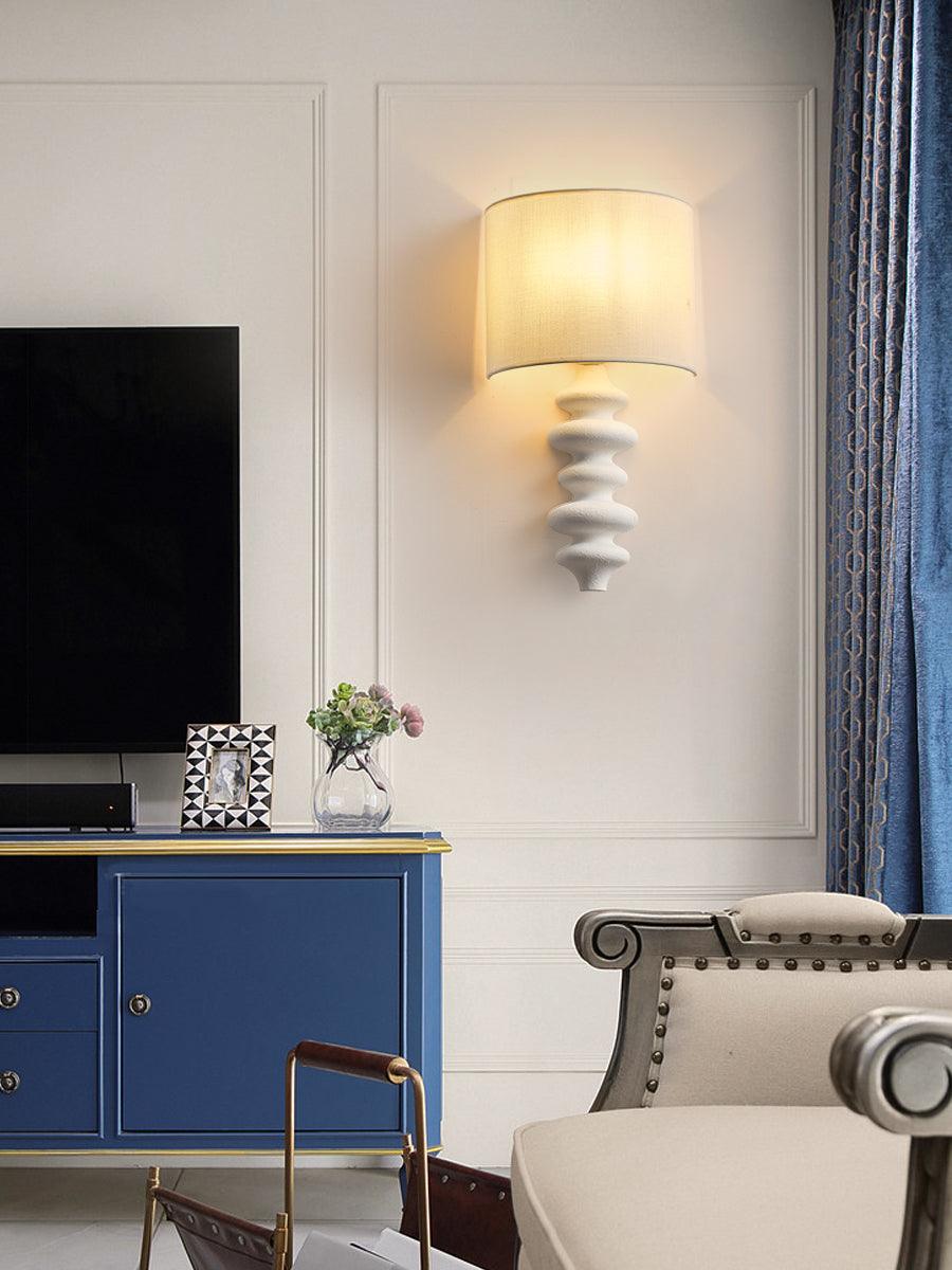 White-column Clérachic wall light, lit, on a panelled wall above a navy sideboard and a television in a living room.