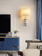 White-column Clérachic wall light, lit, on a panelled wall above a navy sideboard and a television in a living room.
