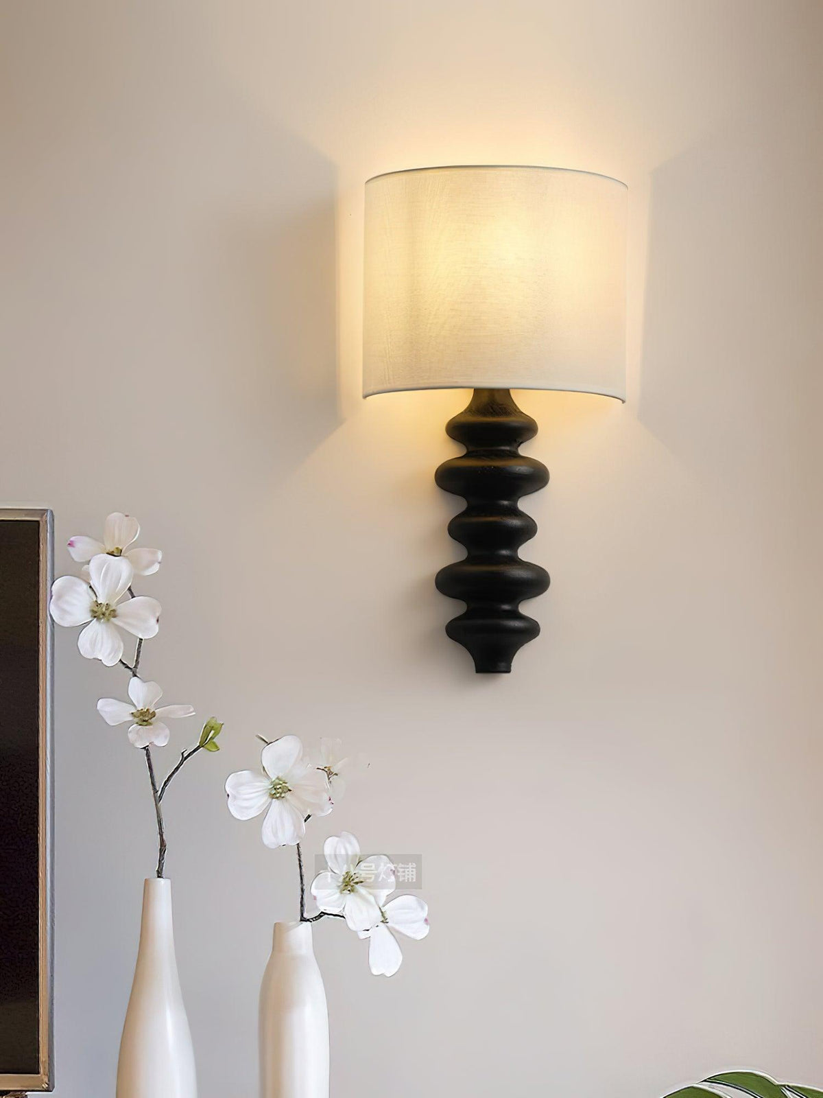 Black-column wall light from Clérachic with a lit white linen shade, above two white vases of blossom on a pale wall.