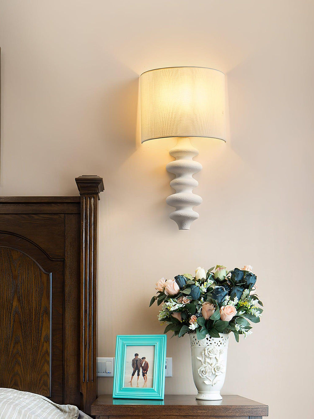 White corrugated Clérachic wall light with a lit linen shade above a bedside table with a teal photo frame and a vase of roses.