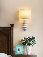 White corrugated Clérachic wall light with a lit linen shade above a bedside table with a teal photo frame and a vase of roses.