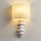 Close view of a lit white-column Clérachic wall light, its linen shade glowing warm above the textured, stacked column.