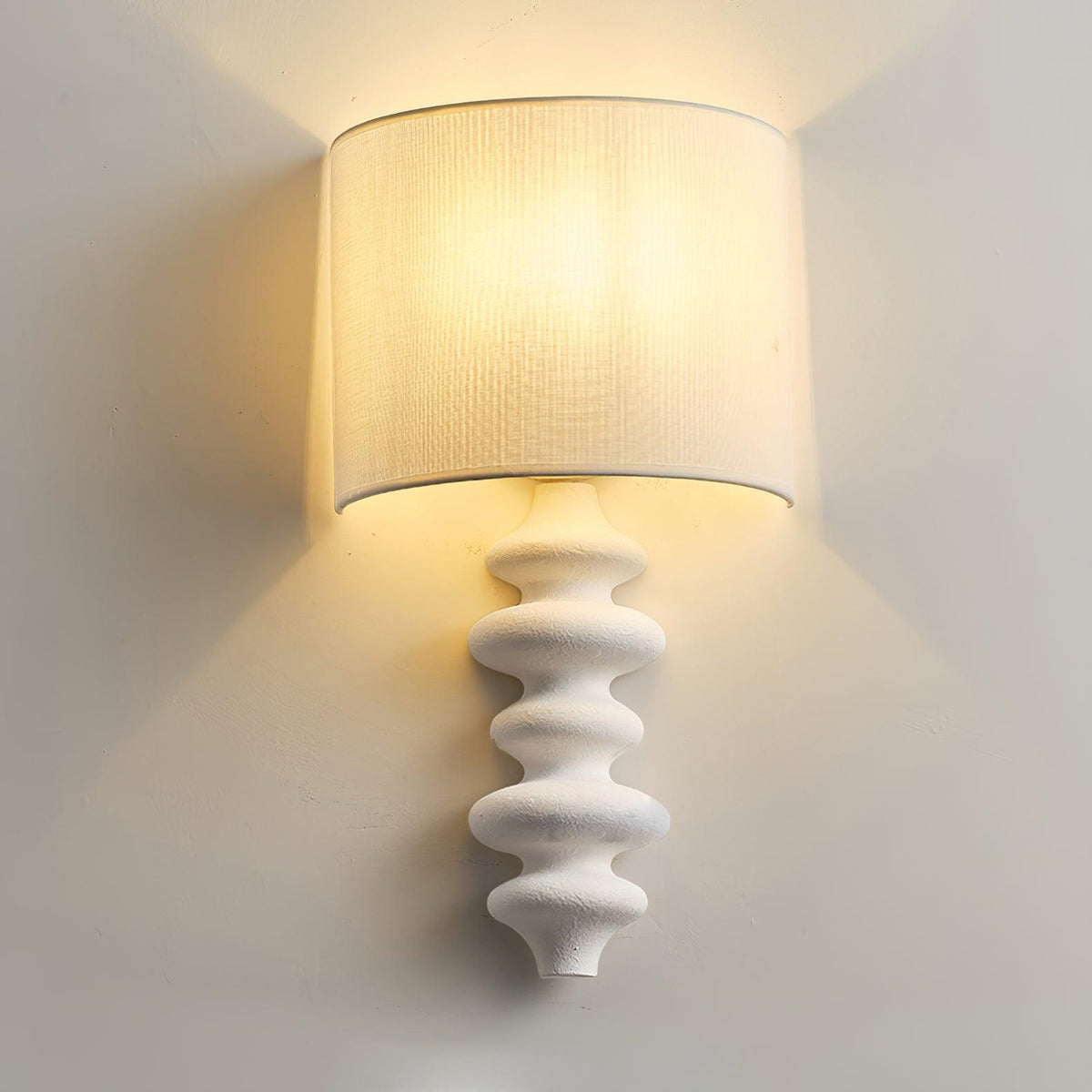 Close view of a lit white-column Clérachic wall light, its linen shade glowing warm above the textured, stacked column.