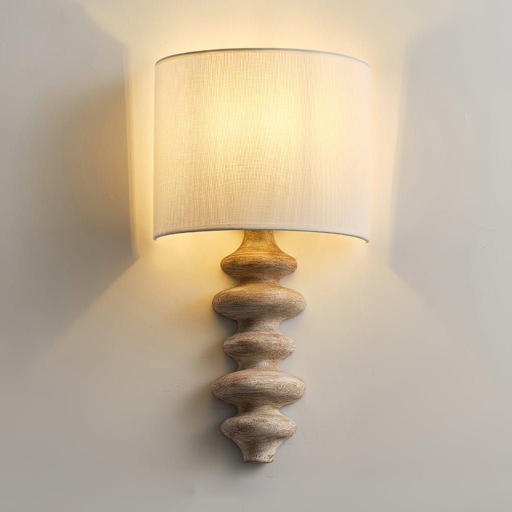 Lit Clérachic wall light with a pale wood column, the linen shade glowing and washing the wall behind it with warm light.