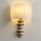 Lit Clérachic wall light with a pale wood column, the linen shade glowing and washing the wall behind it with warm light.