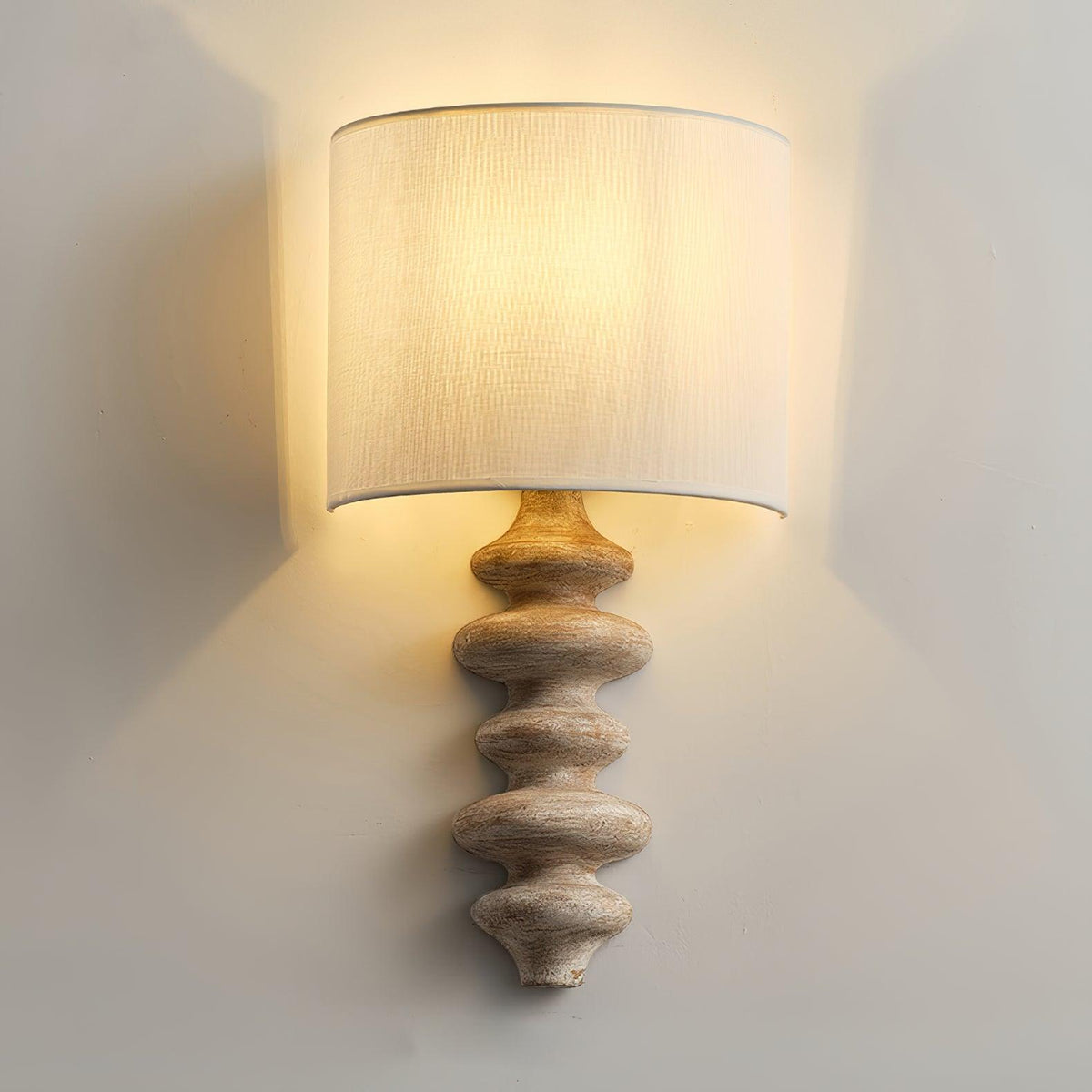 Lit Clérachic wall light with a pale wood column, the linen shade glowing and washing the wall behind it with warm light.