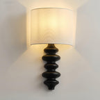 Black-column Clérachic wall light with its white linen shade lit warm, casting light up and down the pale wall.
