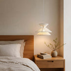 A Clérachic pendant light glows warmly above a wooden bedside table, with a linen-dressed bed and a vase of twigs nearby.