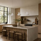 Three white-shaded Clérachic pendant lights over a pale stone kitchen island with dark wood stools and woven seats.
