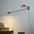 Black and red Clérachic wall light on a pale grey wall, its lamp head casting a soft pool of light above a wooden armchair.