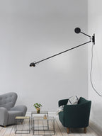 This Clérachic wall light in all black, arm angled down over a grey wall, with a grey armchair and a dark-green sofa below.