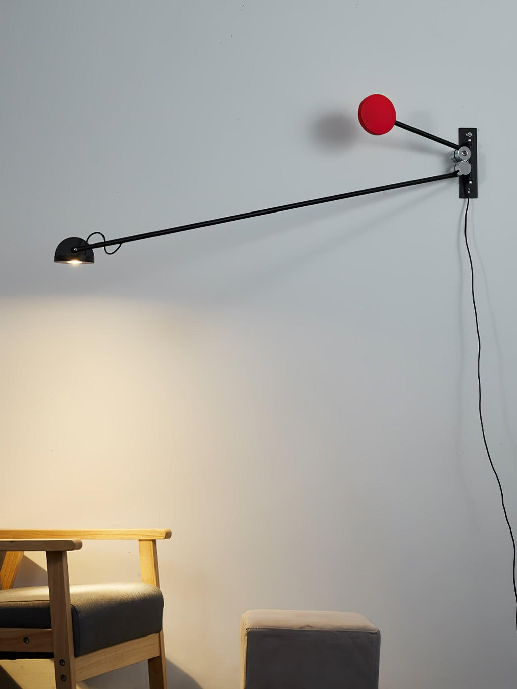 Black and red Clérachic wall light lit above a wooden armchair and grey pouffe, the arm lowered towards the left.