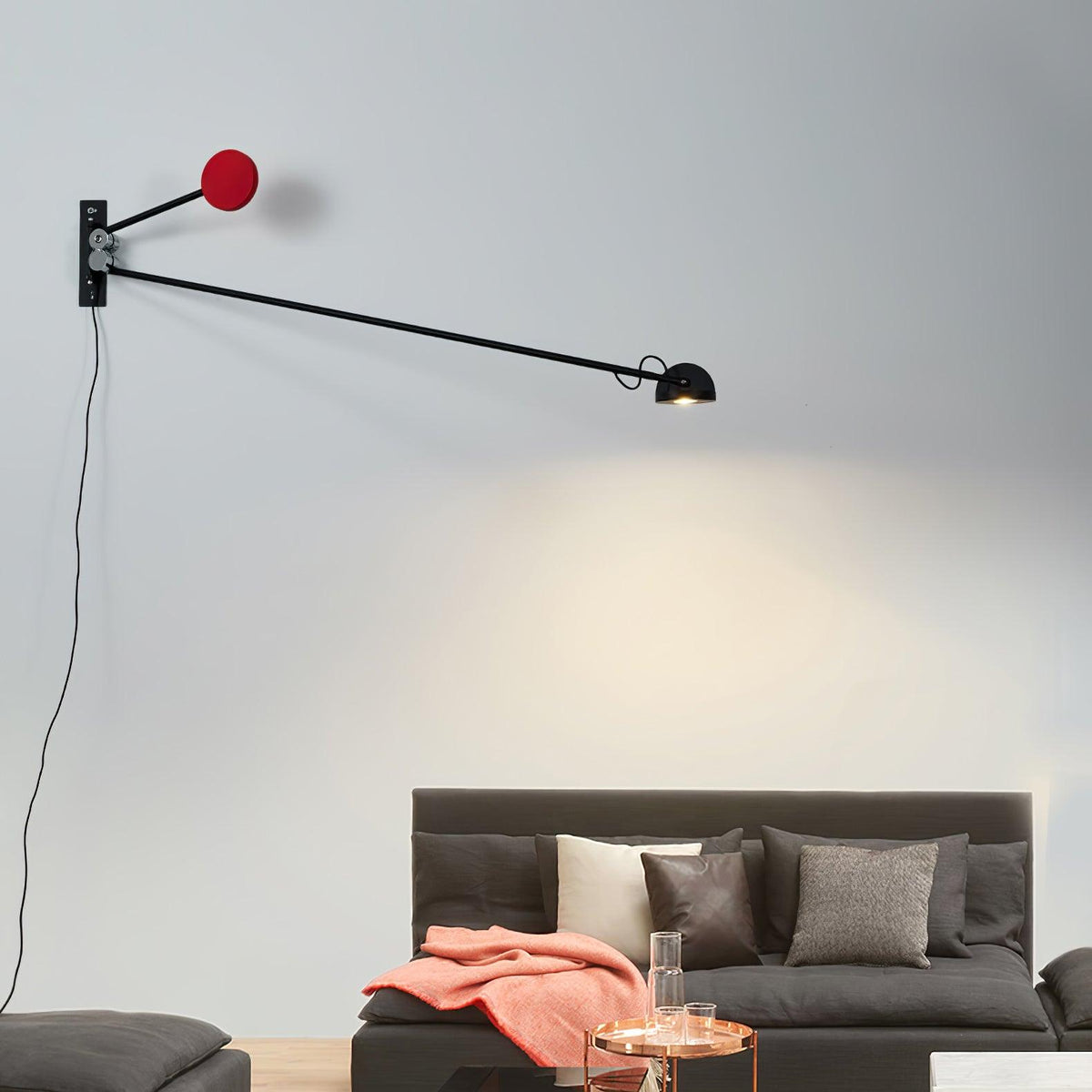 Black and red Clérachic wall light with a long level arm, its lamp head lighting the wall above a charcoal sofa with a coral throw.