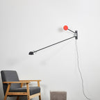 Clérachic wall light with its arm lowered to the left and a red counterweight above, beside a wooden-framed grey armchair.