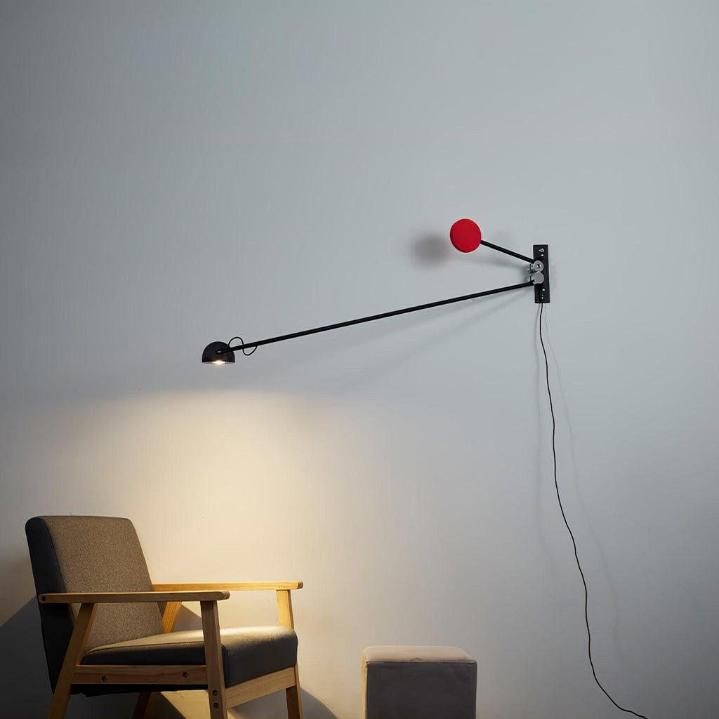 Black and red Clérachic wall light on a blue-grey wall, the lamp head lit above a wooden armchair and a grey pouffe.