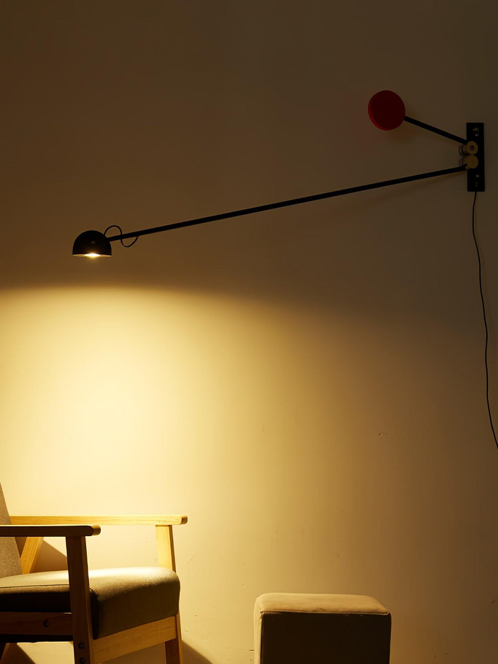A dim, amber-toned room with this Clérachic wall light switched on, the red counterweight above and a wooden armchair in the glow.