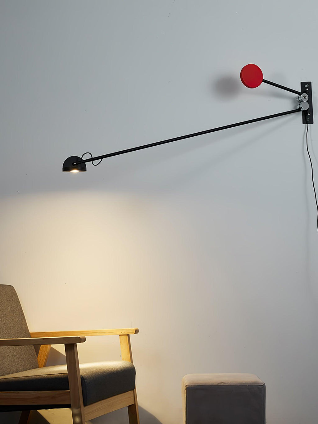 Clérachic wall light with a red counterweight, its arm angled gently down, lighting the wall above a wooden armchair and pouffe.
