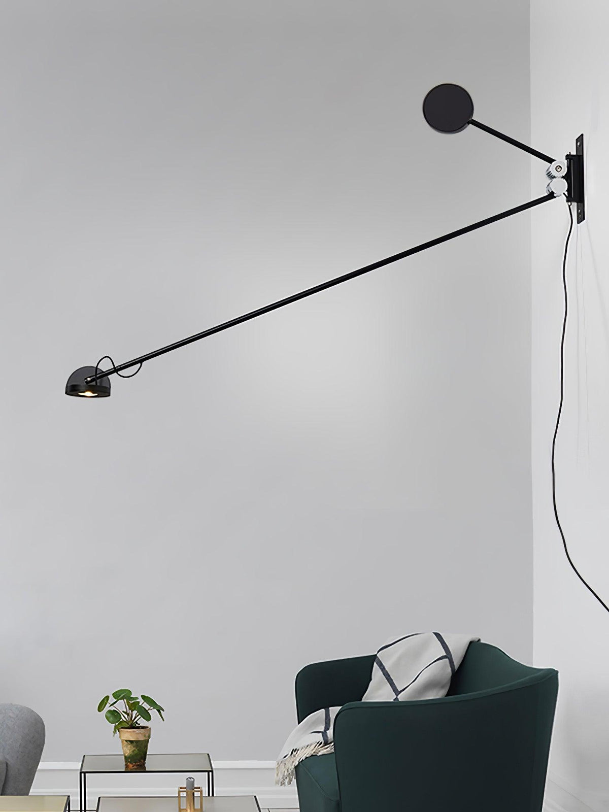Clérachic wall light in all black, the arm angled down from a dark disc counterweight, above a dark-green armchair and a small plant.