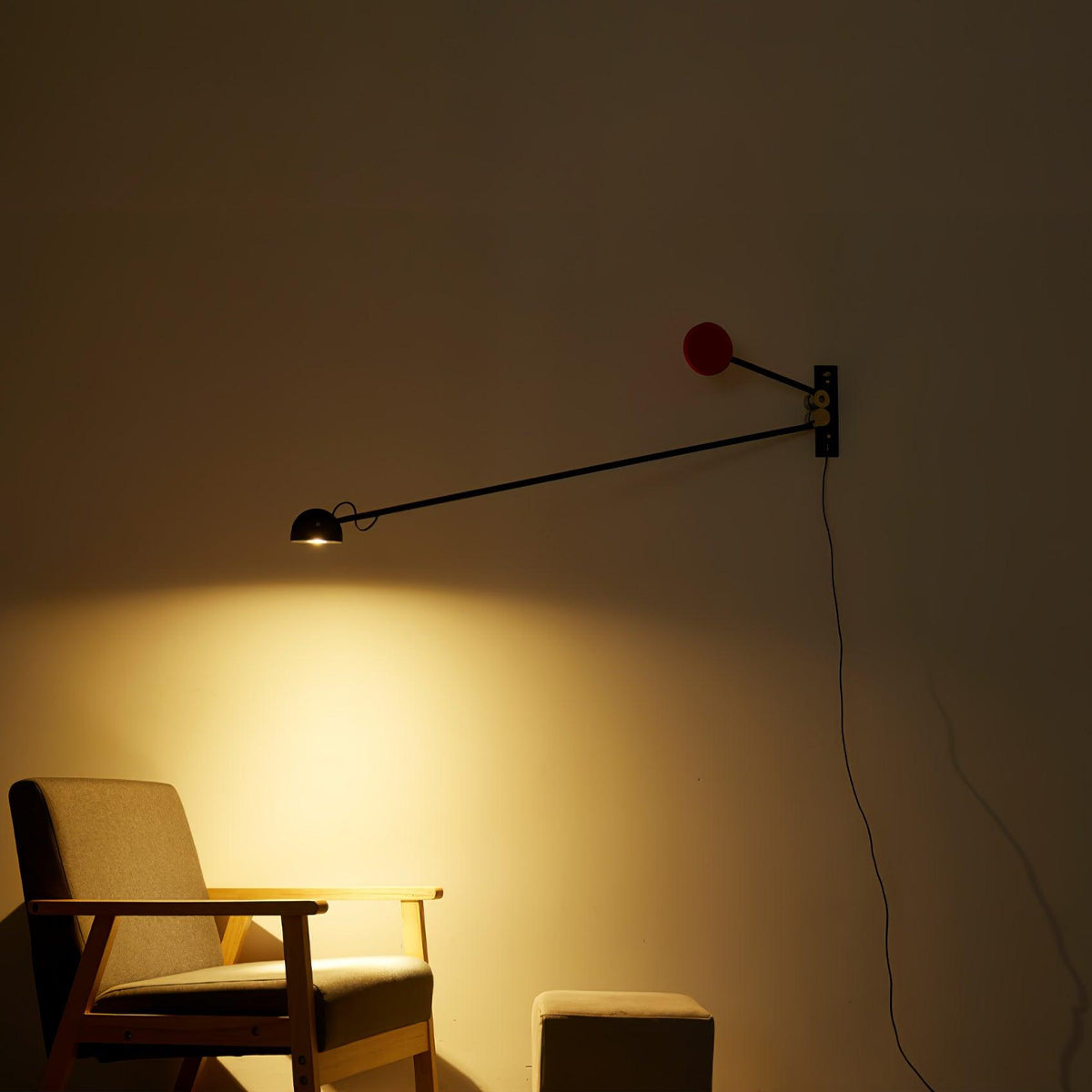 Clérachic wall light lit in a dark room, a bright warm pool of light on the wall beside a wooden armchair, red counterweight above.