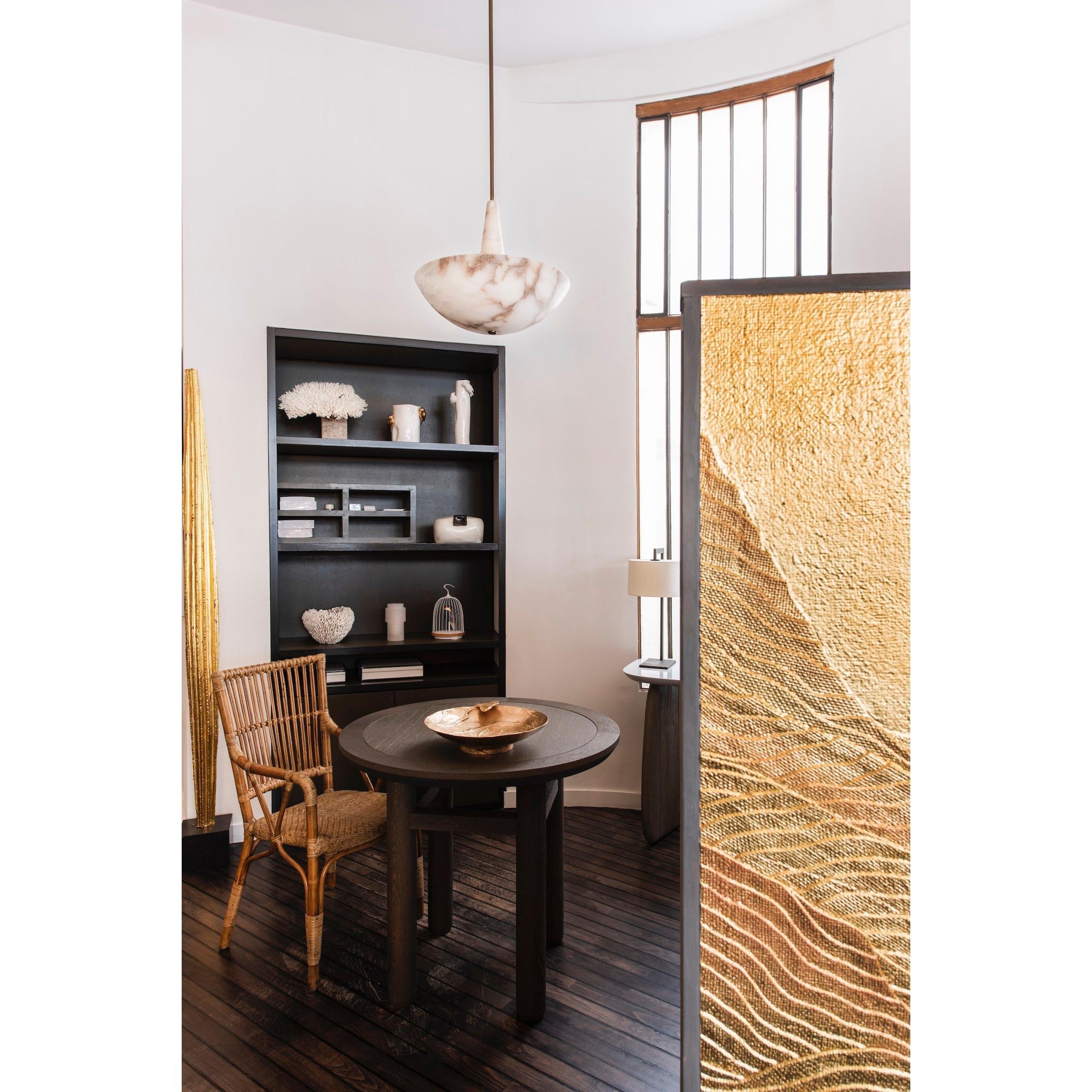 The Clérachic Cove pendant hanging over a round dark-wood table in a corner dining space with rattan chair and gold screen