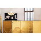 Lit Clérachic Cove alabaster pendant glowing above a black display shelf, seen across a gold folding screen by a tall window