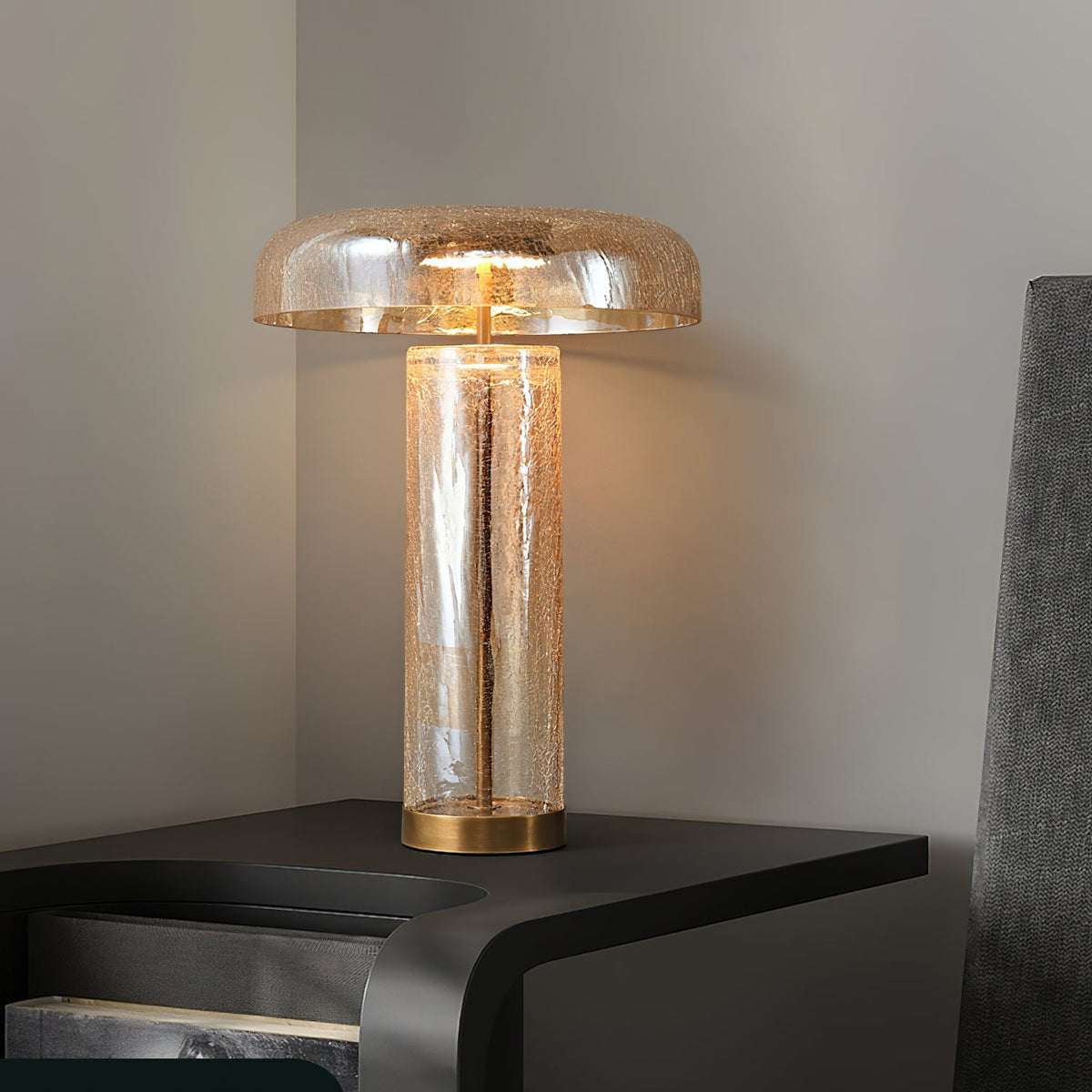 A Clérachic amber crackle-glass table lamp glowing on a dark bedside unit, its domed shade above a clear cylindrical body.