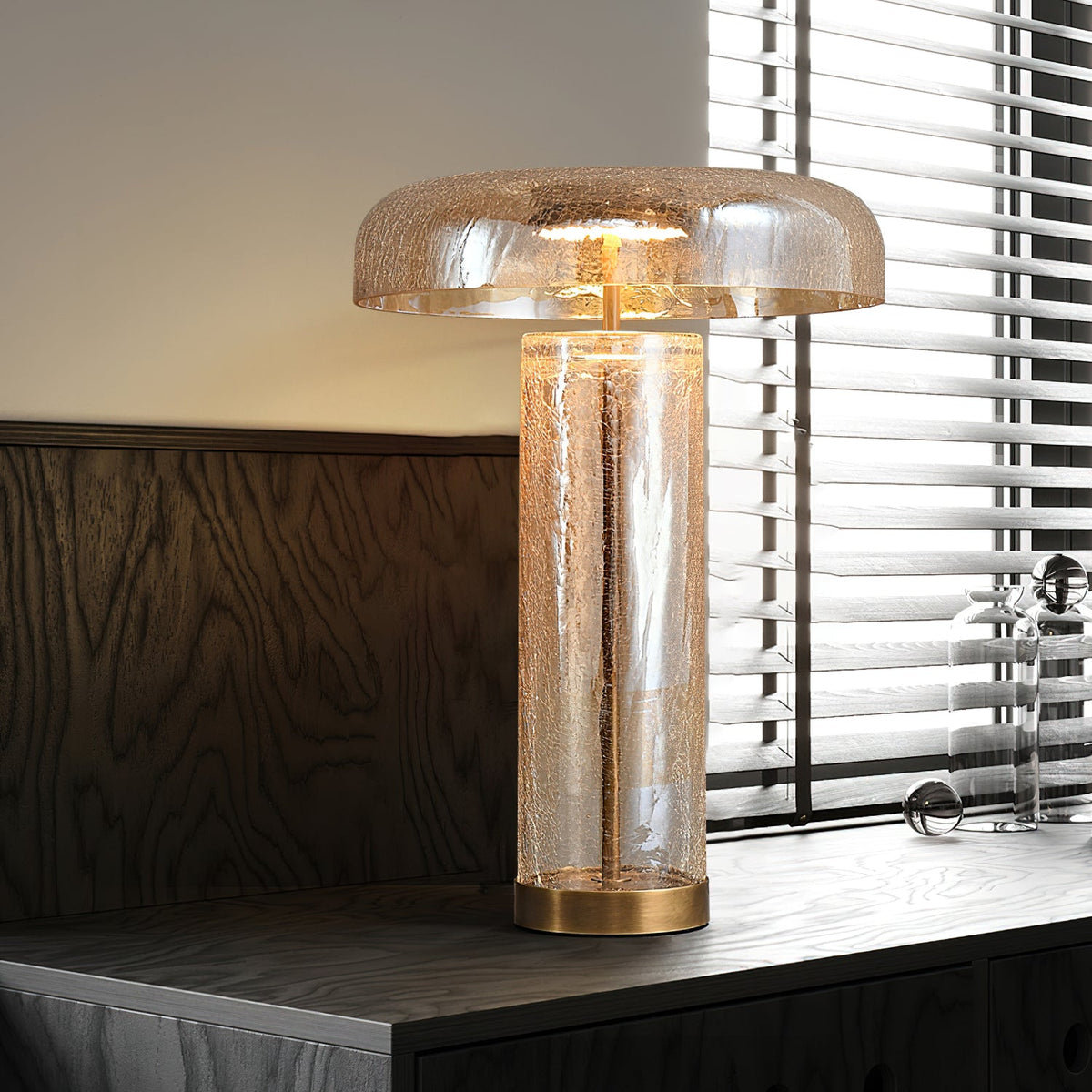 This Clérachic crackle-glass table lamp stands on a dark wood sideboard beside a window with slatted blinds, lit warm.