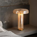 Clérachic amber glass table lamp lit on a pale bedside shelf, against a stone-textured headboard wall with a wall socket.