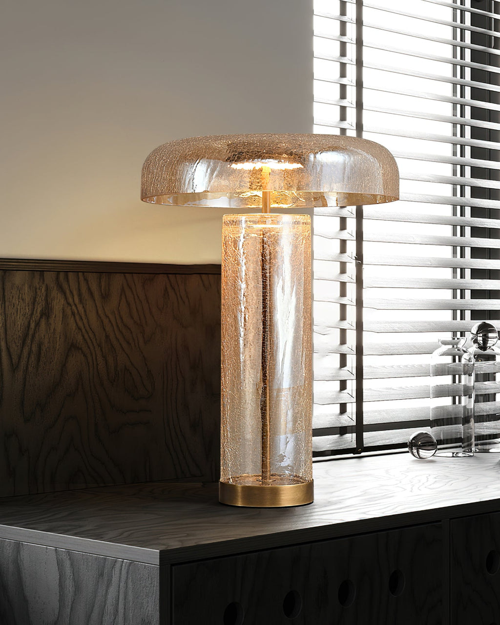 Lamp from Clérachic on a dark wood cabinet below a blind-covered window, the cracked glass body warm with light.