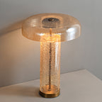 Lit Clérachic table lamp on a grey surface, the amber dome glowing above a crackled clear glass body with a bronze base.