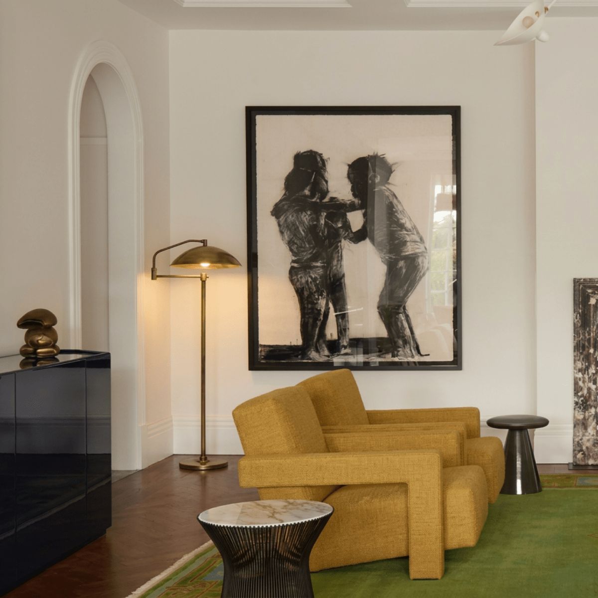 Clérachic brass floor lamp lit beside mustard armchairs, under a large framed charcoal drawing, on a green rug.