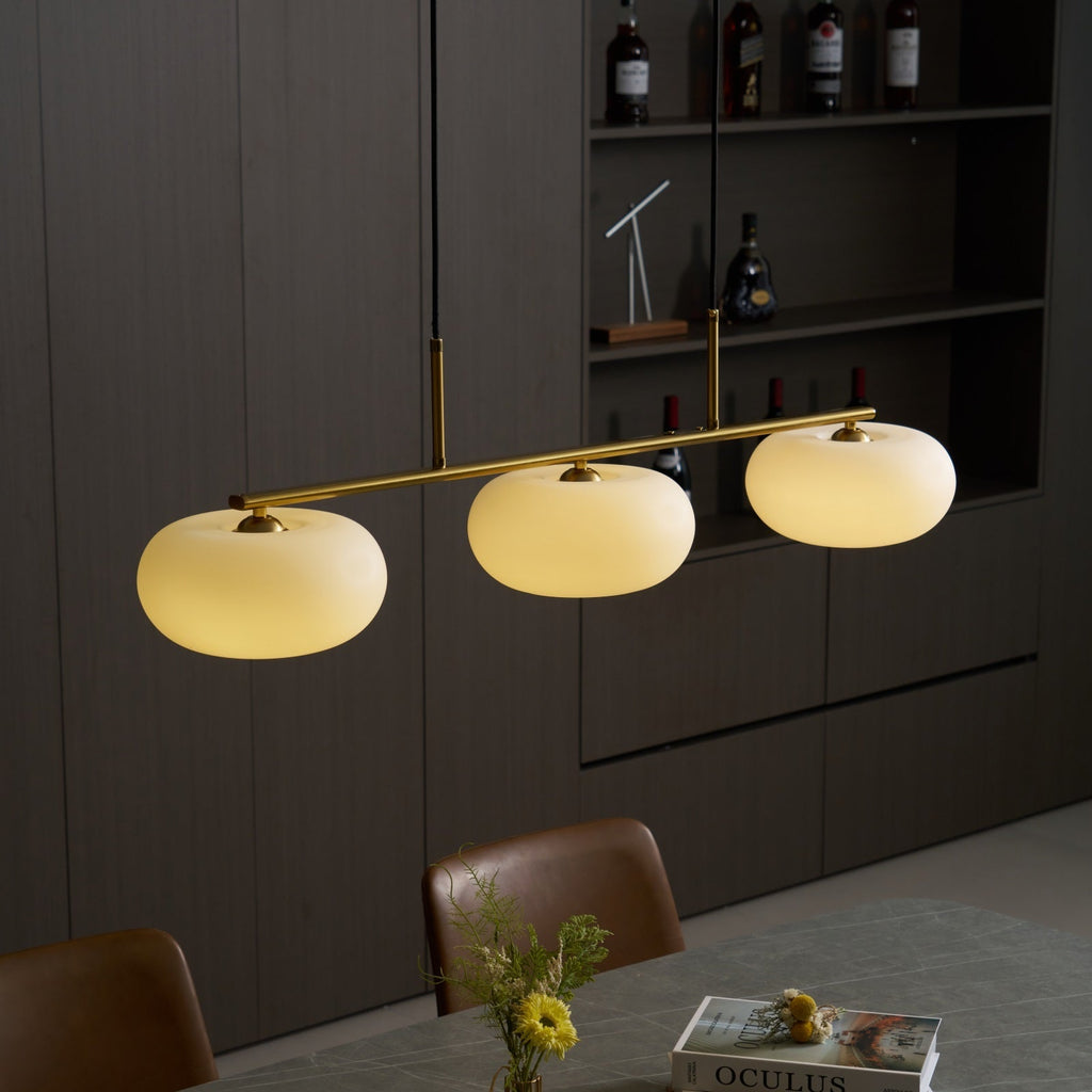 Clérachic cream glass pendant light in gold, glowing over a grey dining table with an OCULUS book and yellow flower.