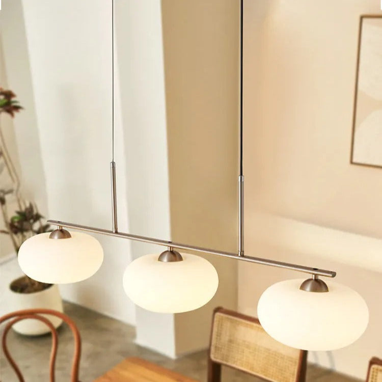 Clérachic gold-frame cream glass pendant over a wood dining table, two gold rods and a rattan chair behind.