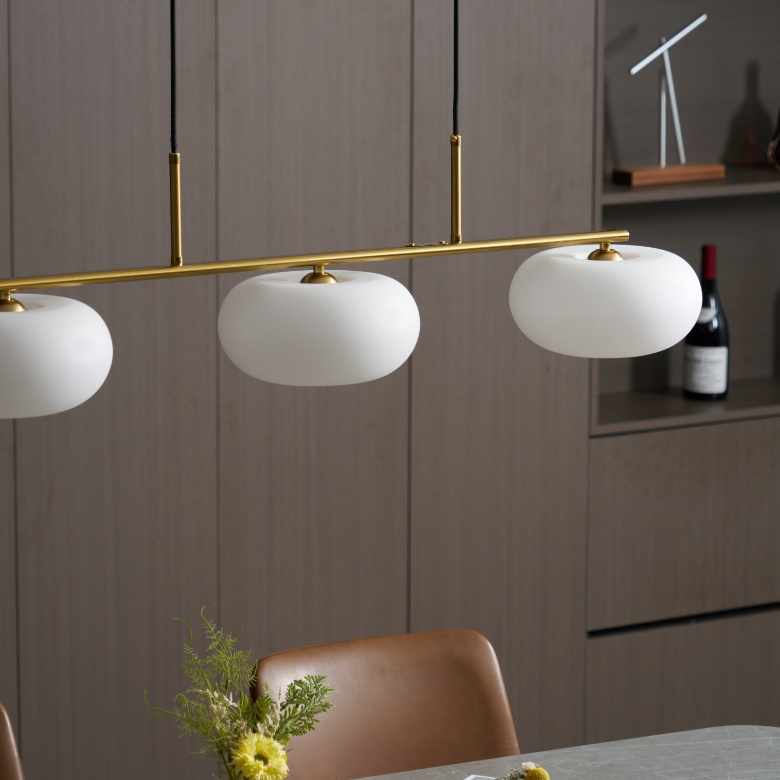 Clérachic gold-frame cream glass pendant over a dining table, brown leather chair and yellow flowers in front.