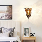 Warm light from a Clérachic wall sconce in amber glass falls across a cream wall above a bed with striped cushions and a pale nightstand.