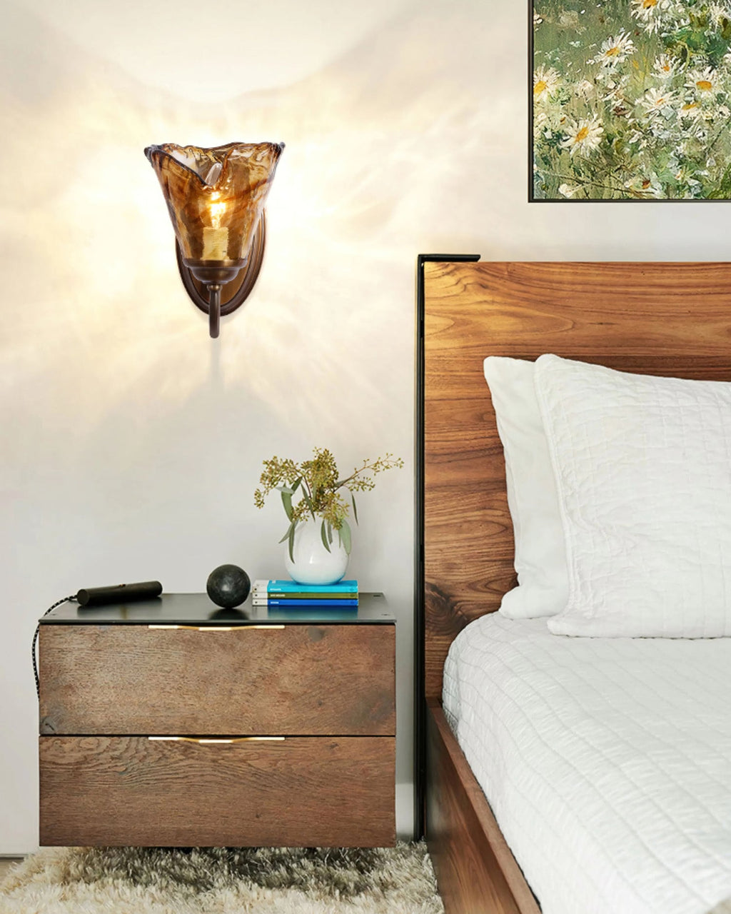 Tall crop of a Clérachic amber glass wall sconce above a dark wood bedside table, with white bedding at the right.