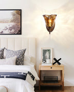 Wide view of a bedroom with a Clérachic amber glass sconce on the wall, a bed with a blanket and a woven shelf beside it.