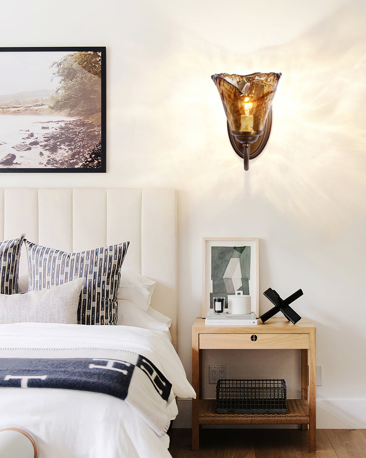Wide view of a bedroom with a Clérachic amber glass sconce on the wall, a bed with a blanket and a woven shelf beside it.