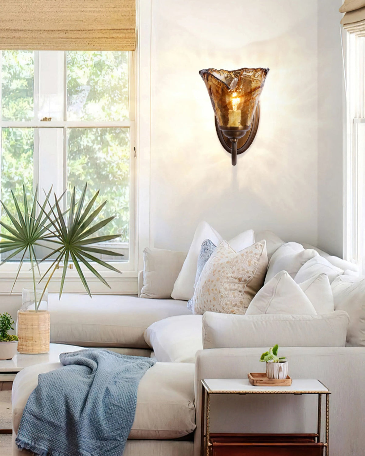 In a bright living room with a palm plant and cream corner sofa, a Clérachic amber glass wall sconce glows on a white wall.