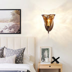Close crop of a bedroom wall where a Clérachic amber glass sconce glows above a nightstand, with a framed print to the left.