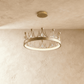 Clérachic crown-shaped gold ring chandelier with kite-cut crystals and gold ball finials, lit from below against a limewash wall.