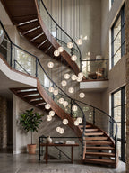 A long fall of glowing Clérachic crystal balls through a double-height stairwell, beside a wooden spiral stair and black metal rail.
